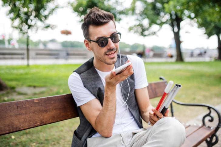 Young blind man with smartphone sitting on bench in park in city, calling