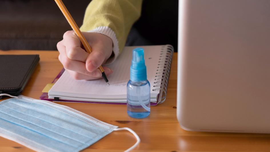 student writing notes with mask and hand sanitizer on table